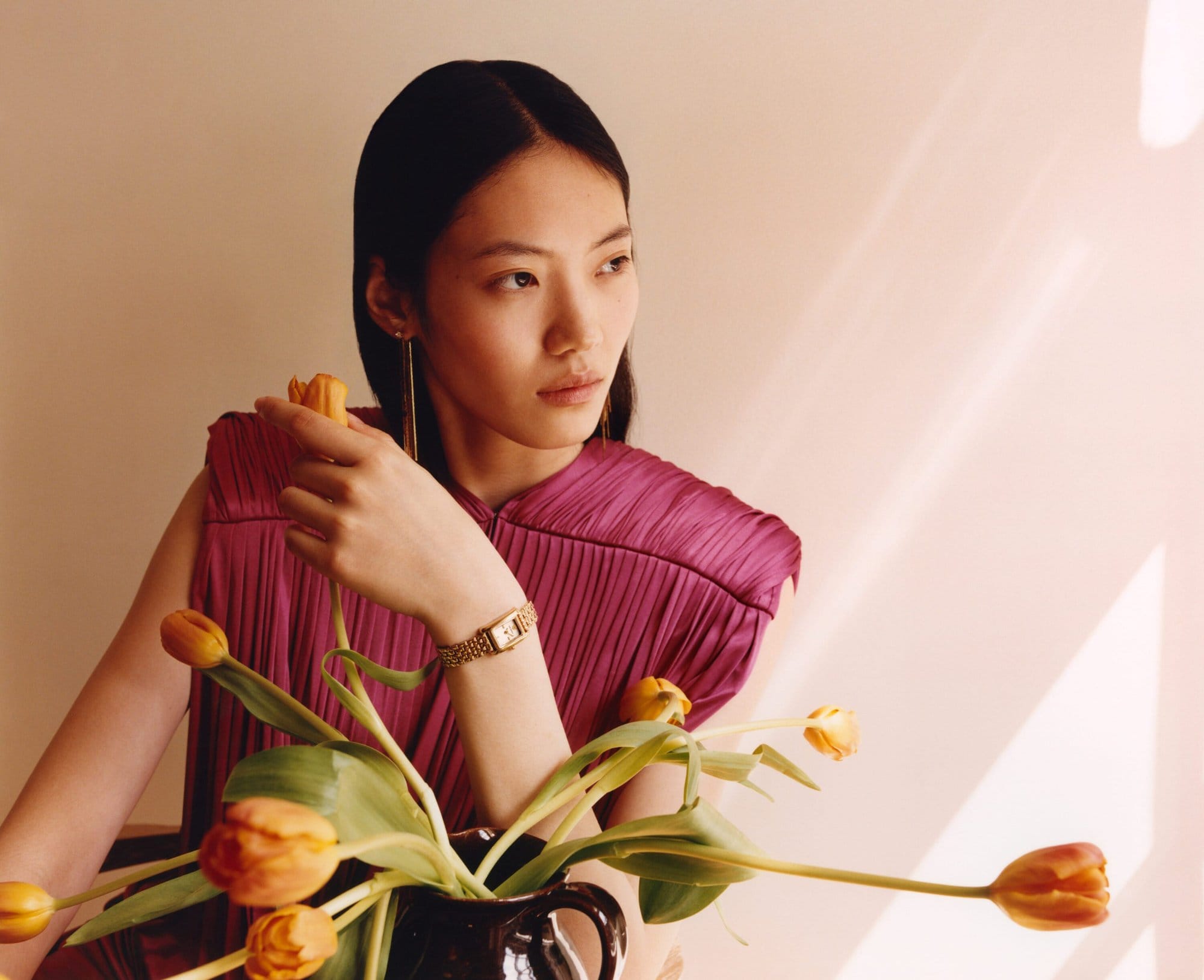 East Asian female model in magenta pleated top holding an orange tulip near a dark vase with flowers
