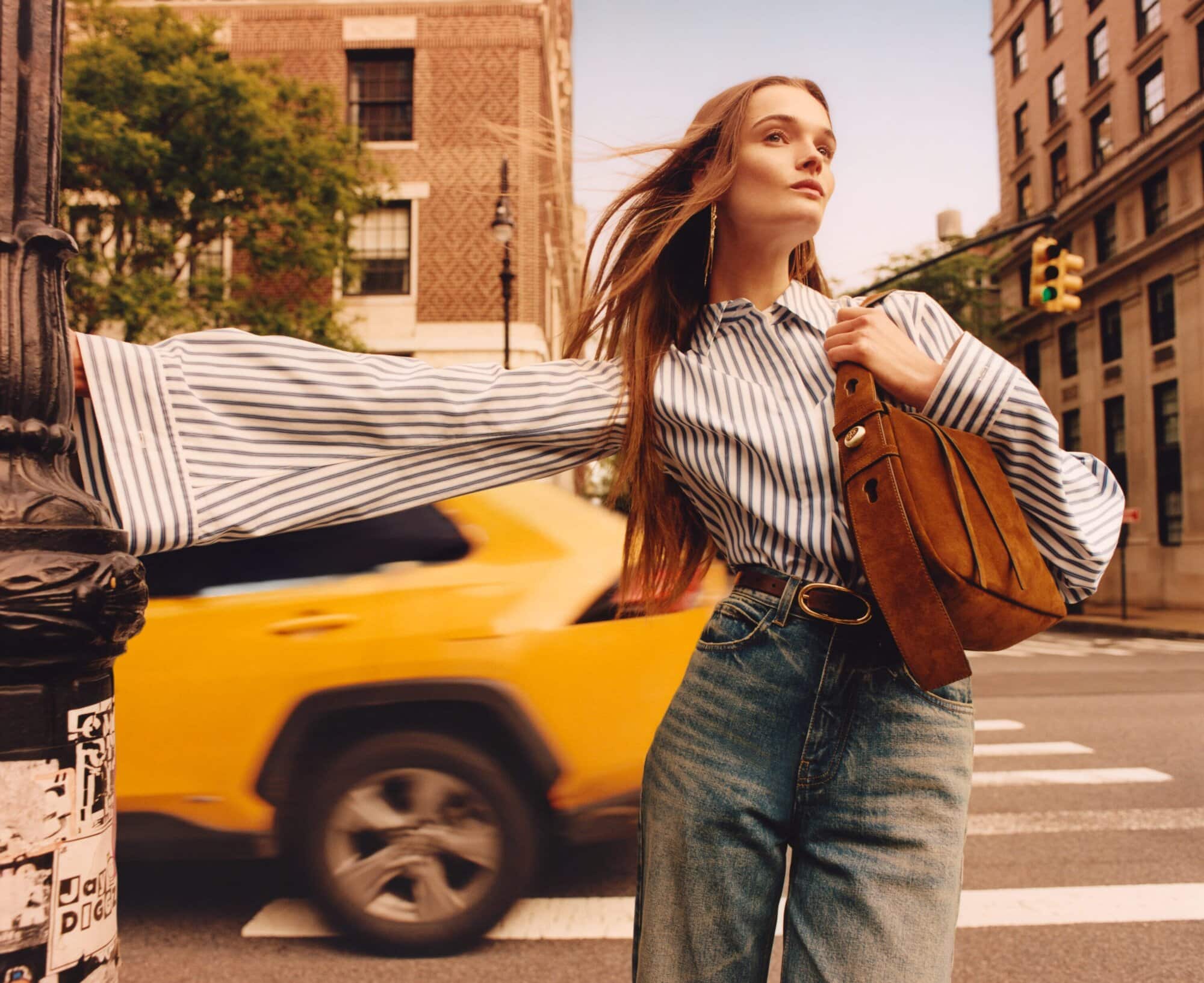 Model in blue-and-white striped shirt and blue jeans leaning against a post in New York City with a yellow taxi in the background