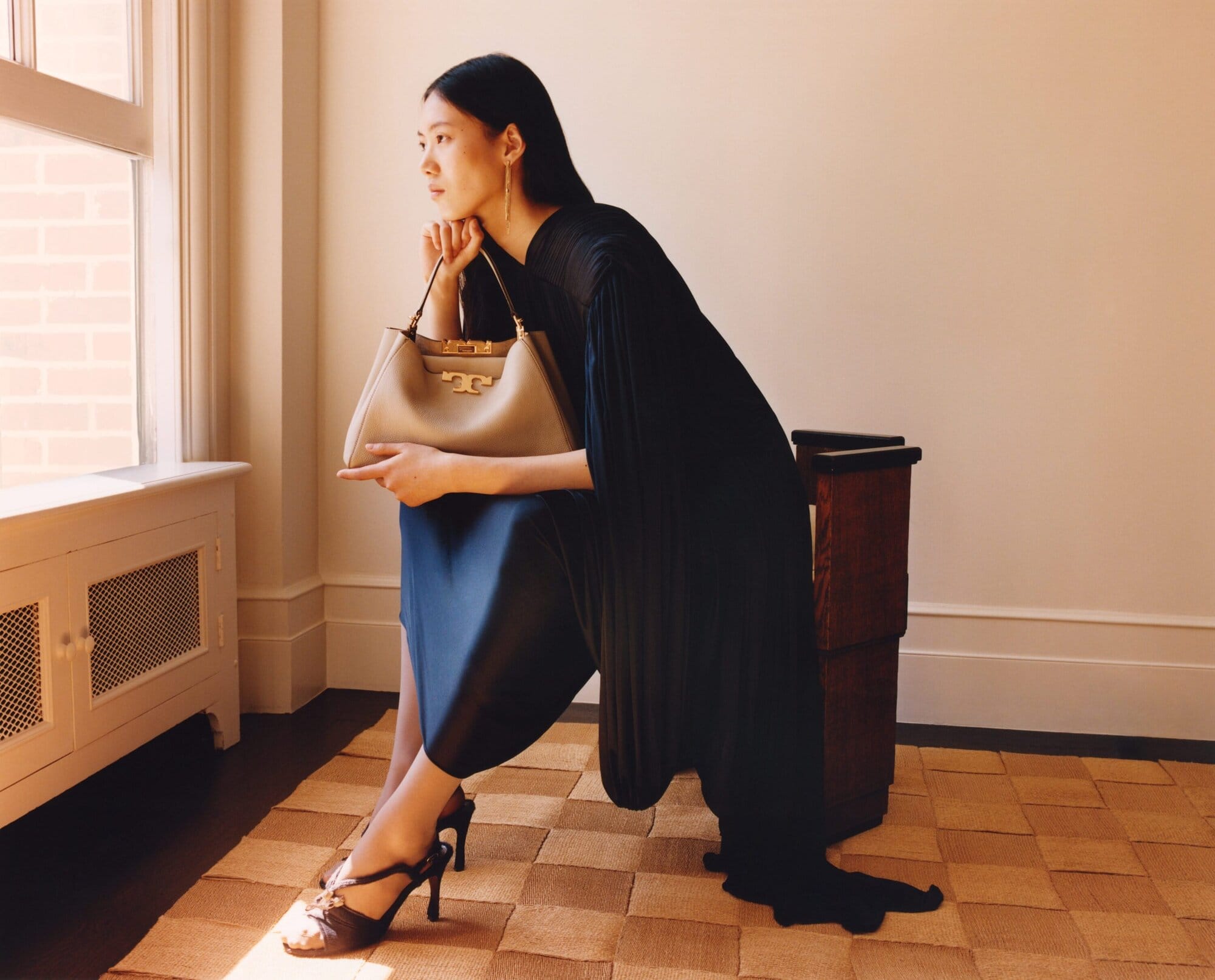 Model wearing dark dress seated indoors near a window holding a light-colored handbag for Tory Burch Fall 2025 campaign.