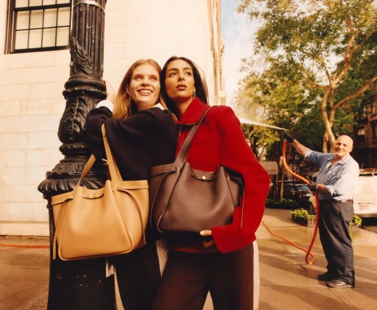 Two women holding leather tote bags pose against a lamppost on a New York City street during daylight.
