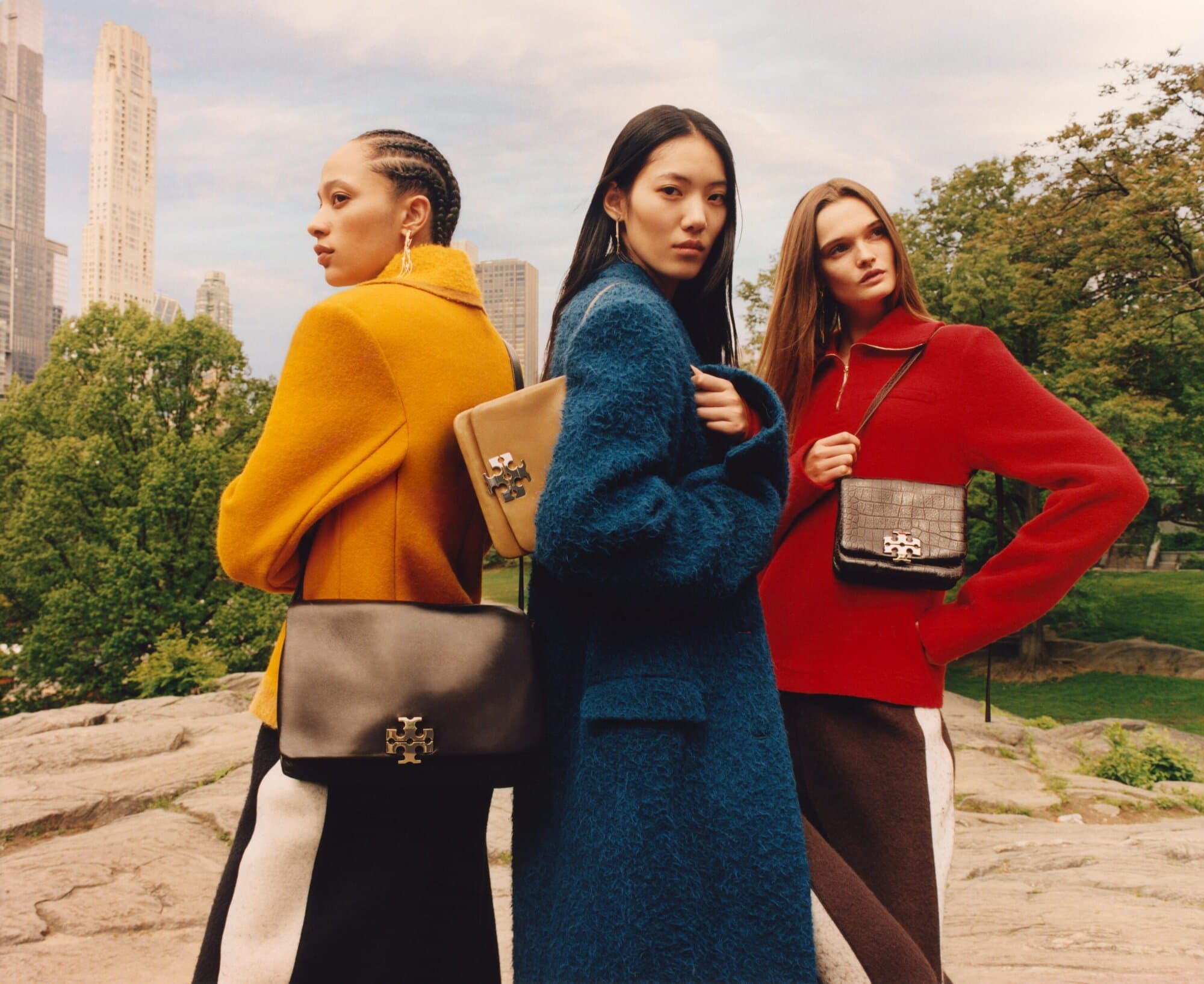 Three female models posing outdoors in New York City wearing brightly coloured coats and carrying designer handbags.