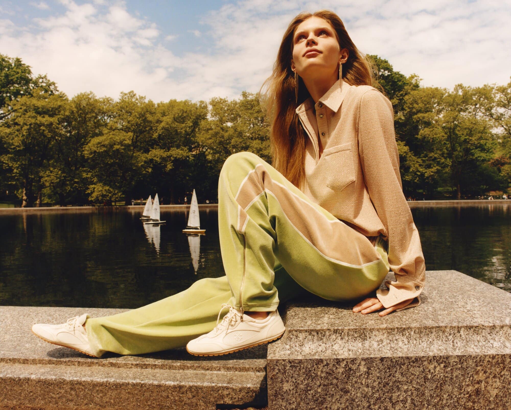 Model seated on a stone wall by a lake wearing a beige collared top and green trousers in New York City