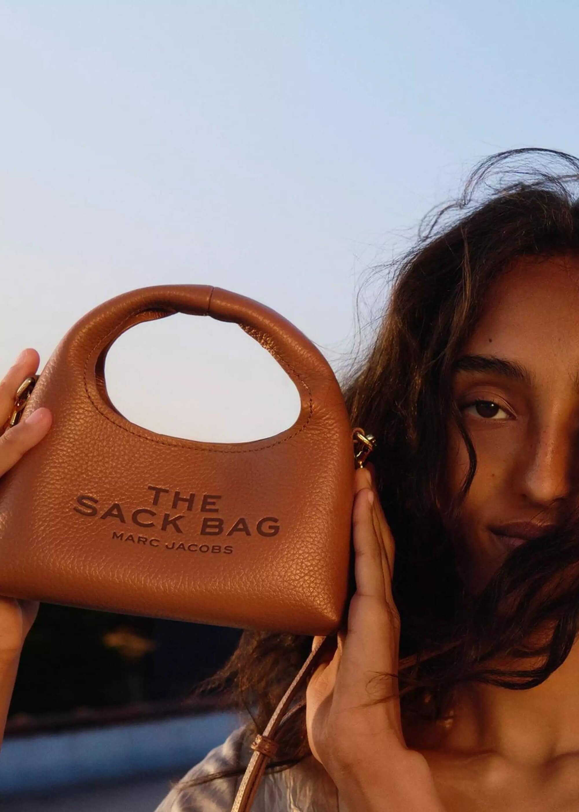 Close-up portrait of model holding a brown leather Marc Jacobs handbag against an outdoor sky background