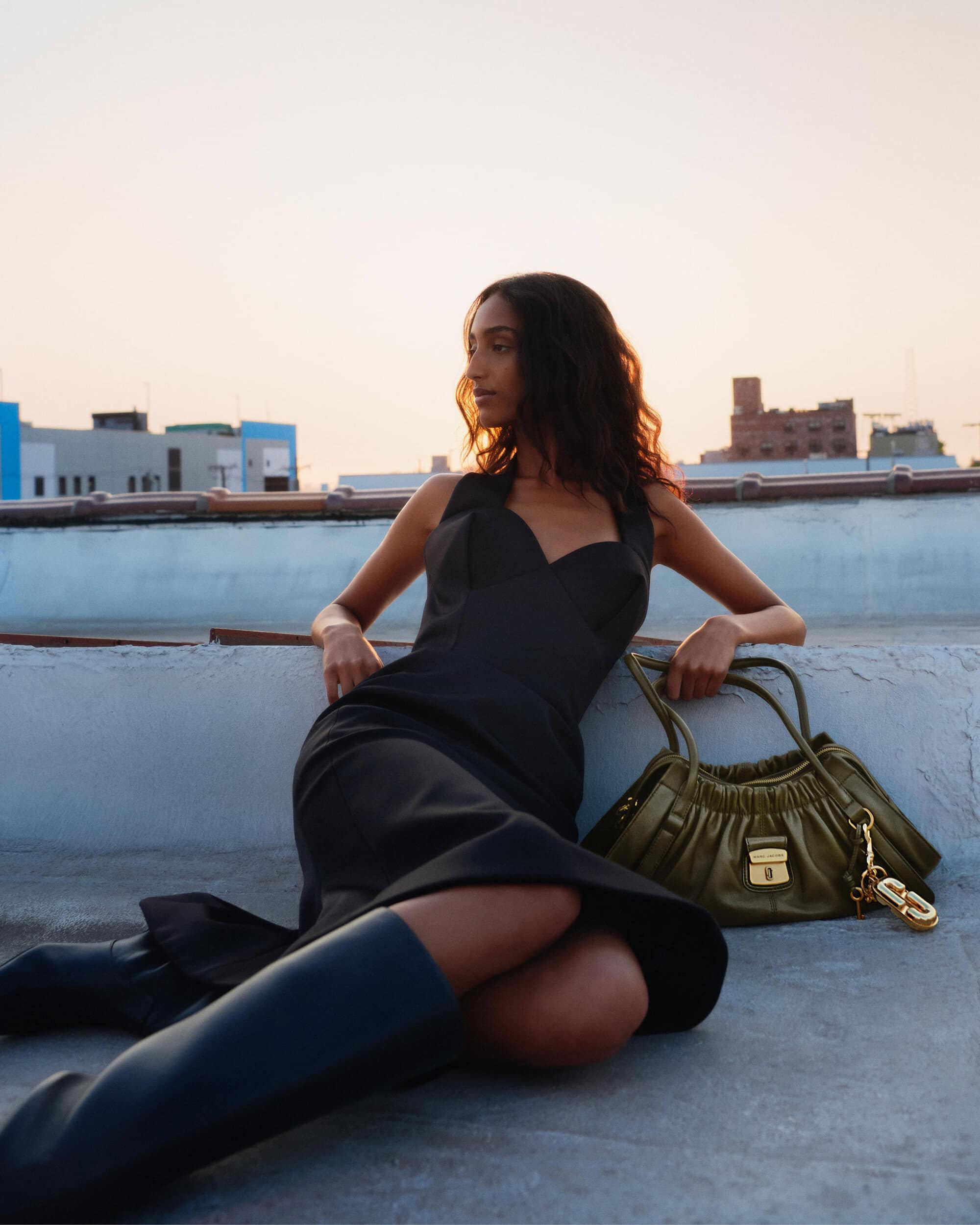 Model wearing a black dress and dark boots leaning on a rooftop ledge with an olive green handbag beside her.