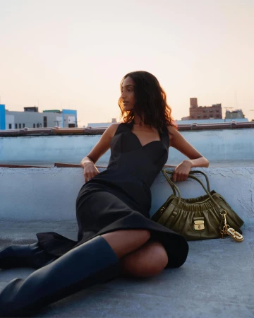 Model wearing a black dress and dark boots leaning on a rooftop ledge with an olive green handbag beside her.