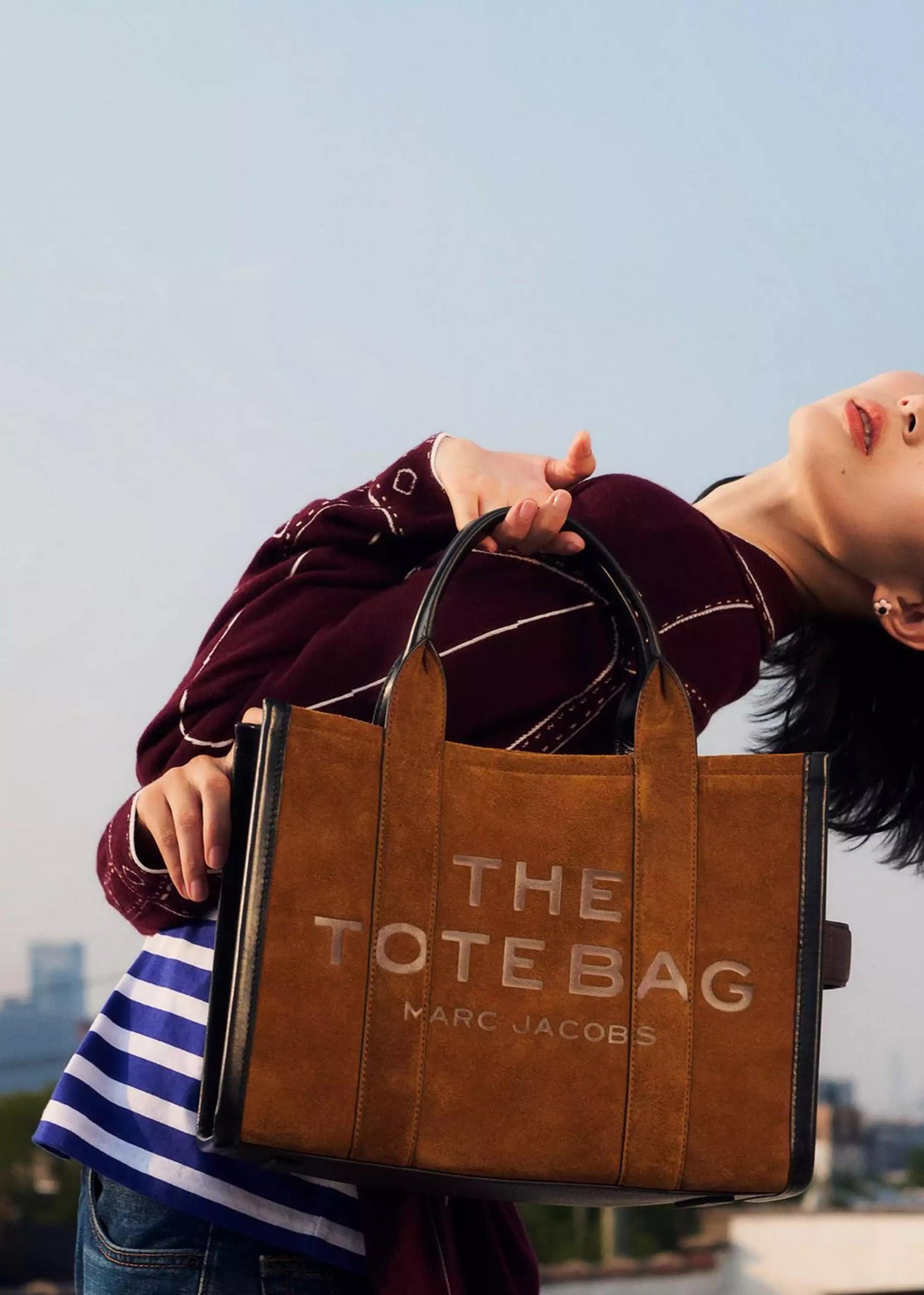 Model holding a brown suede tote bag in front of a blurred urban background