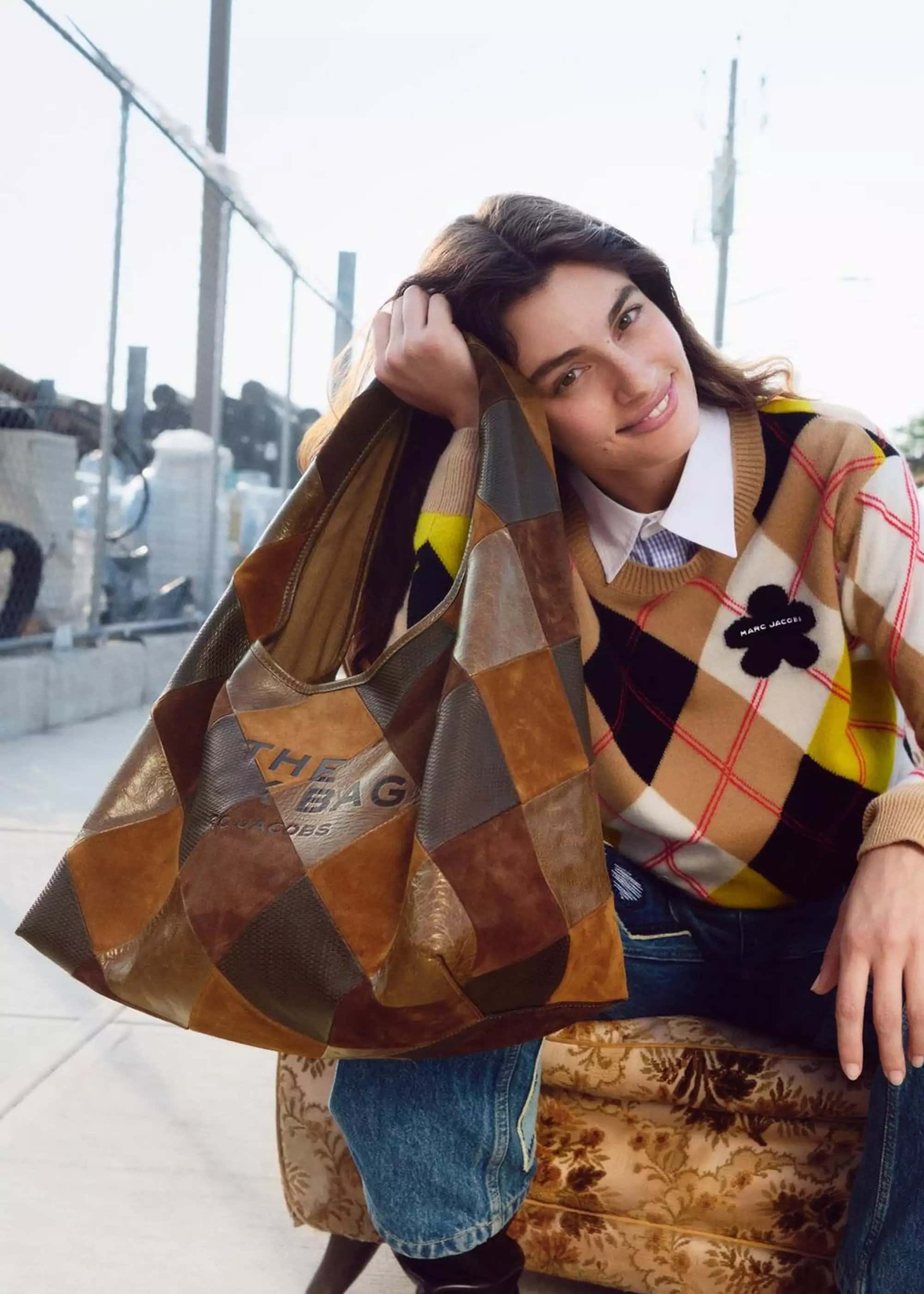 Model holding a large patchwork leather hobo bag wearing an argyle sweater and jeans, outdoors against chain-link fence