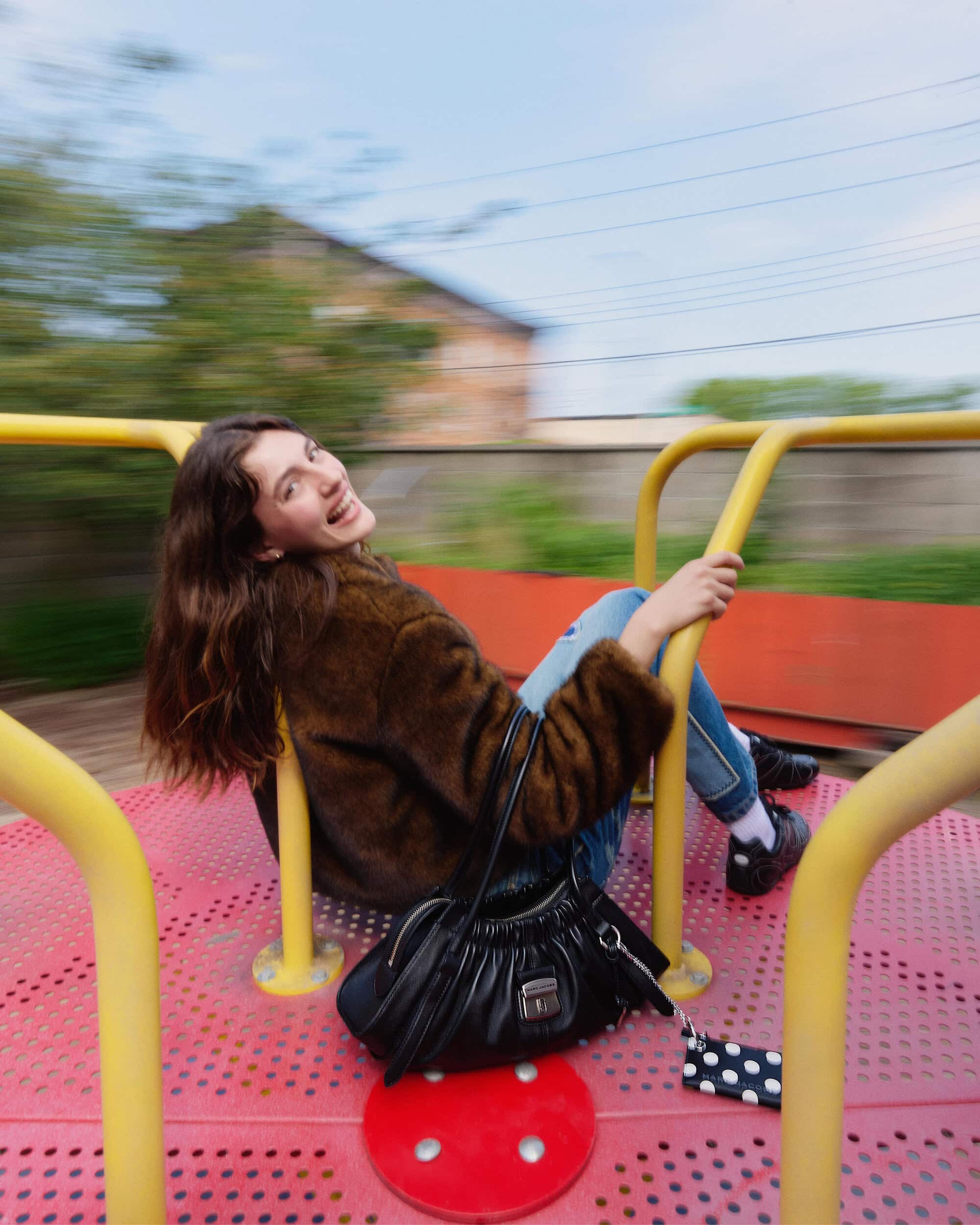 Woman in brown furry jacket riding a spinning playground merry-go-round holding a black handbag.