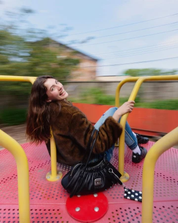 Woman in brown furry jacket riding a spinning playground merry-go-round holding a black handbag.