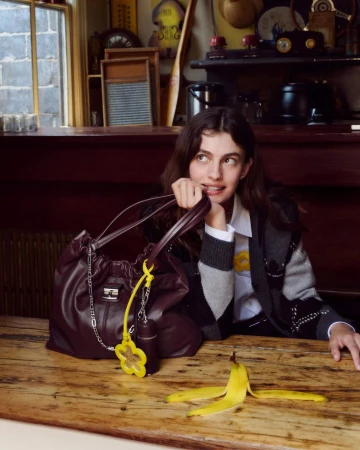 Model seated at wooden table with a large dark leather handbag and a banana peel, captured indoors.