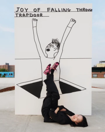 A person lying on a rooftop beneath a large hand-drawn art panel featuring a figure falling through a trapdoor.