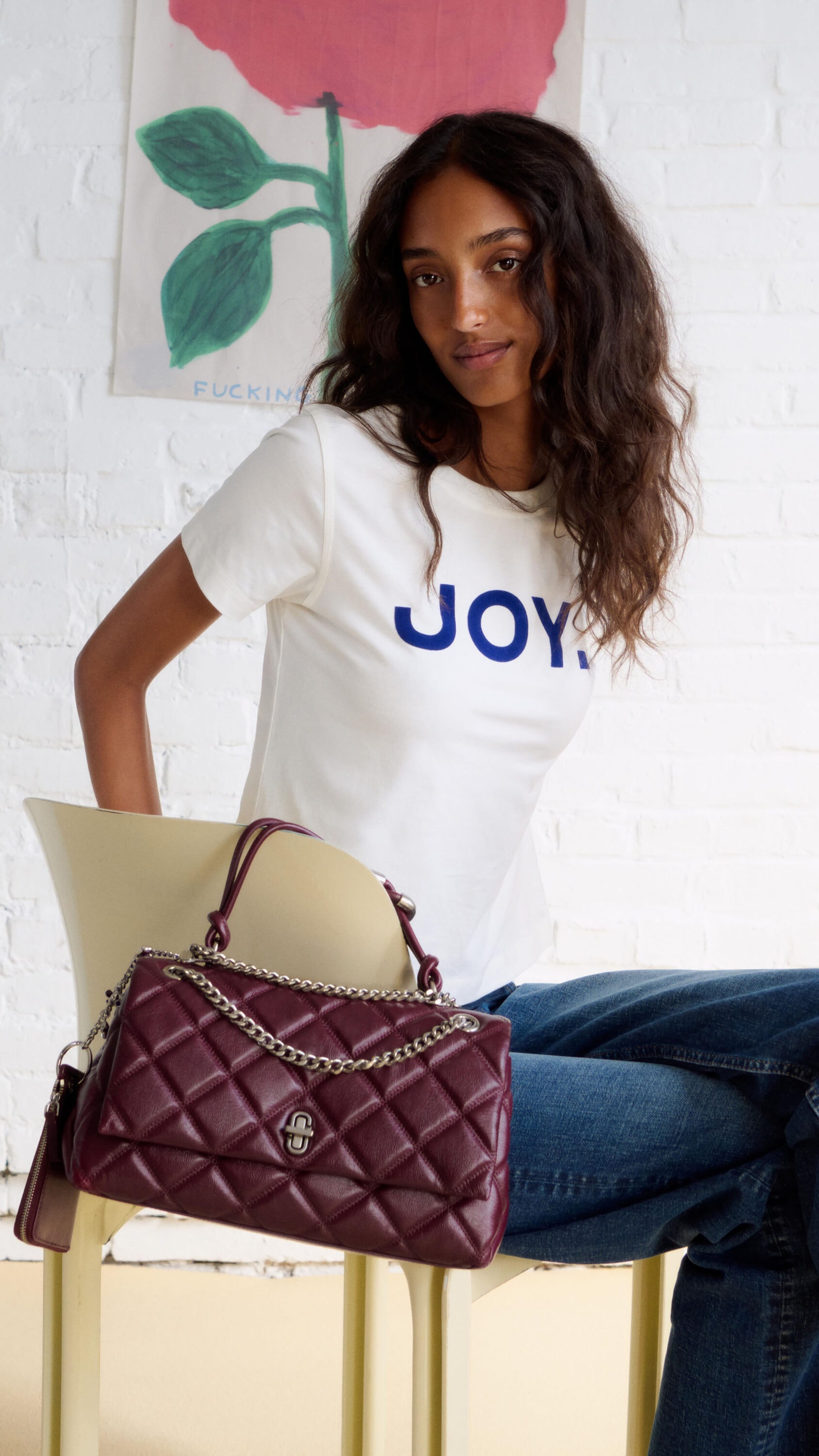 Model Mona Tougaard wearing white Joy t-shirt and blue jeans, seated on yellow chair holding a quilted burgundy leather handbag