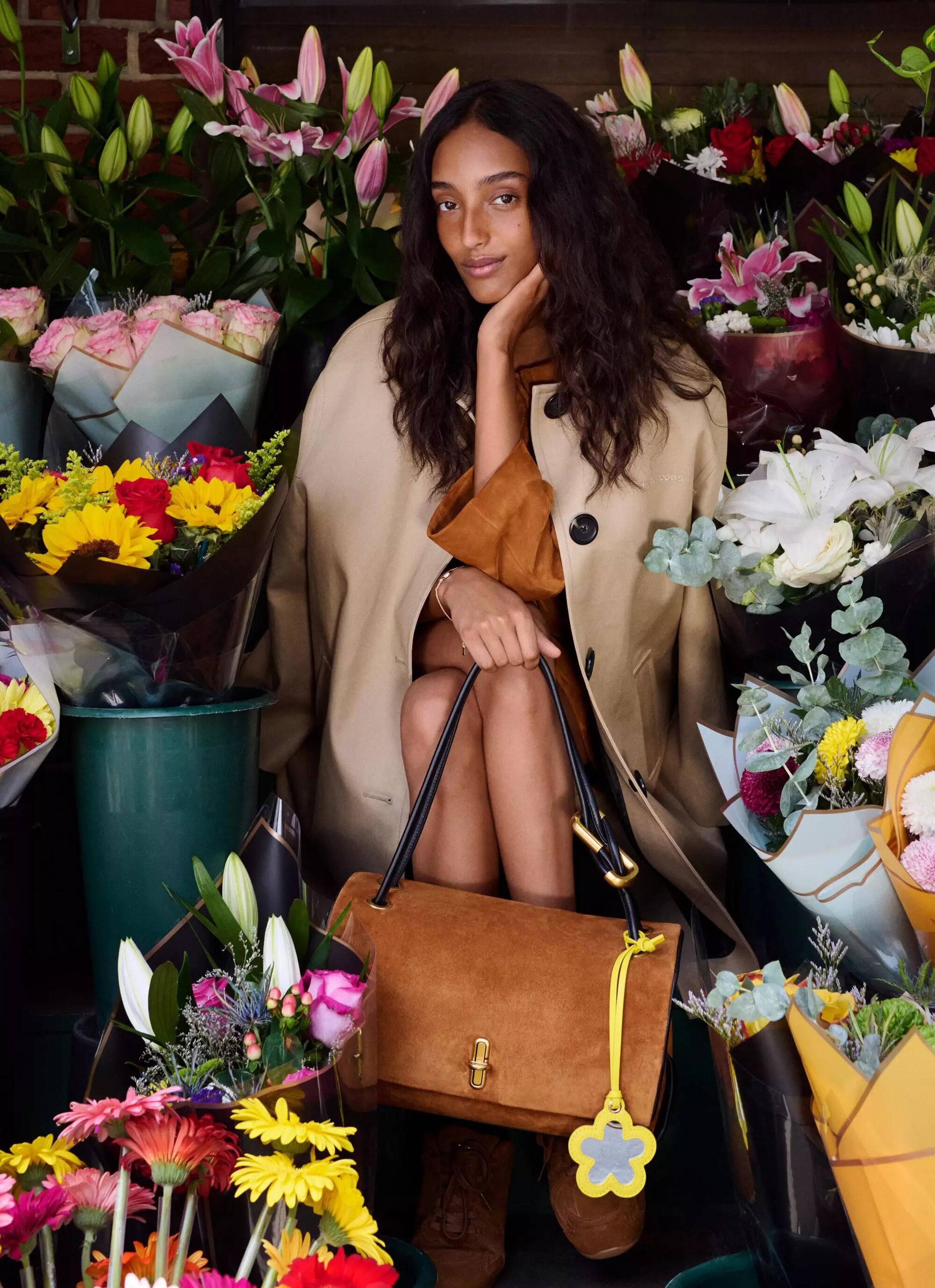 Model seated among floral bouquets holding a brown suede handbag for Marc Jacobs