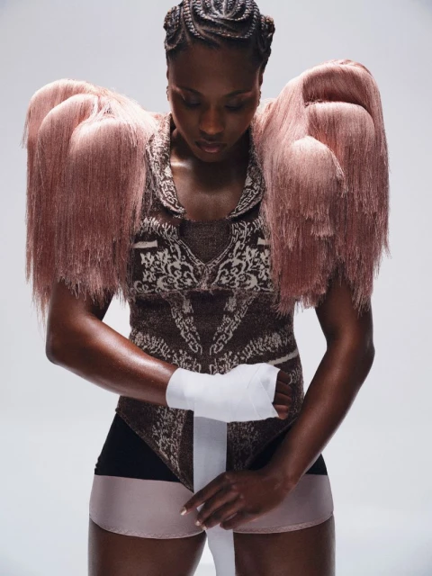 Fashion shot of a Black female adult with braids wrapping her hand with white bandage, wearing a patterned bodysuit and fringed shoulder pads.