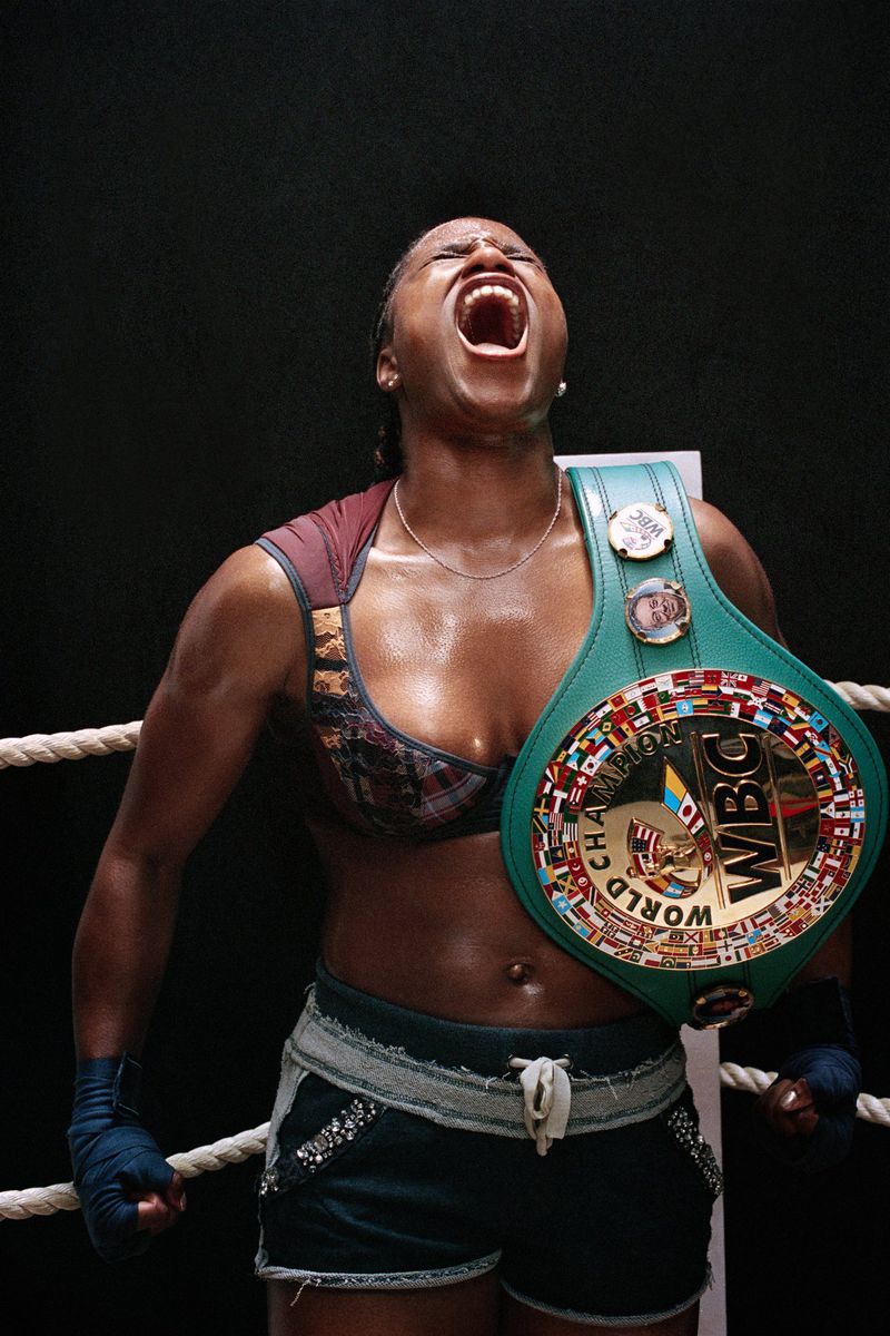 Female boxer celebrates victory, wearing a championship belt and boxing wraps, with a dark background and bright lighting.