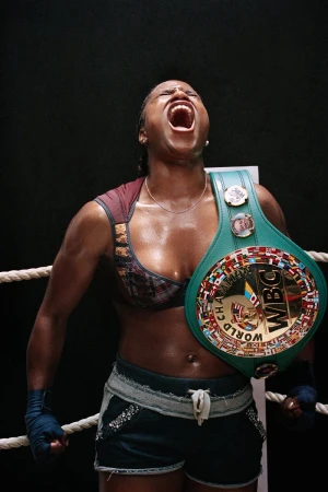 Female boxer celebrates victory, wearing a championship belt and boxing wraps, with a dark background and bright lighting.