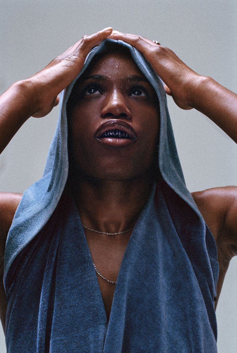 Portrait of an African American woman in a hooded sleeveless top looking upward, hands near her head, against a light background.