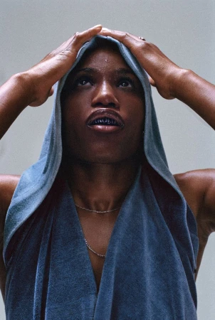 Portrait of an African American woman in a hooded sleeveless top looking upward, hands near her head, against a light background.
