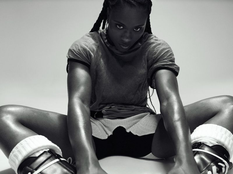 Athletic dark-skinned woman with braided hair, wearing workout clothes and retro sneakers, is sitting with legs apart against a white backdrop.