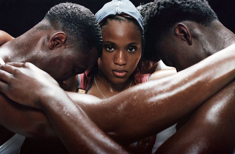 Close-up of determined African-American woman embraced by two male athletes in a dimly lit room, showcasing sweat and intense focus.