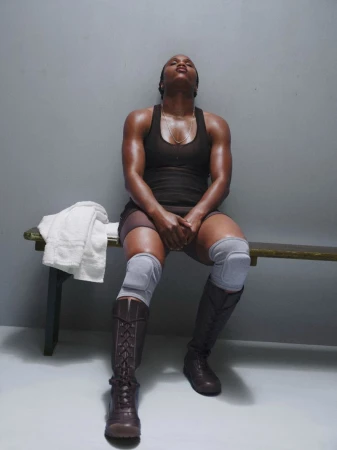 Athletic African-American woman resting indoors on a bench wearing knee pads and boots, after a workout, looking up towards the light.