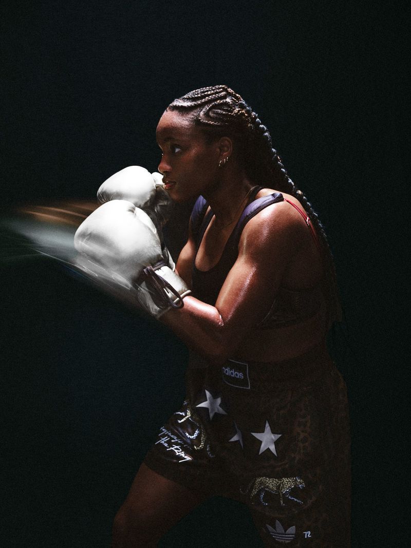 Strong female boxer with braided hair in motion wearing gloves and patterned shorts against a dark background in a studio setting.