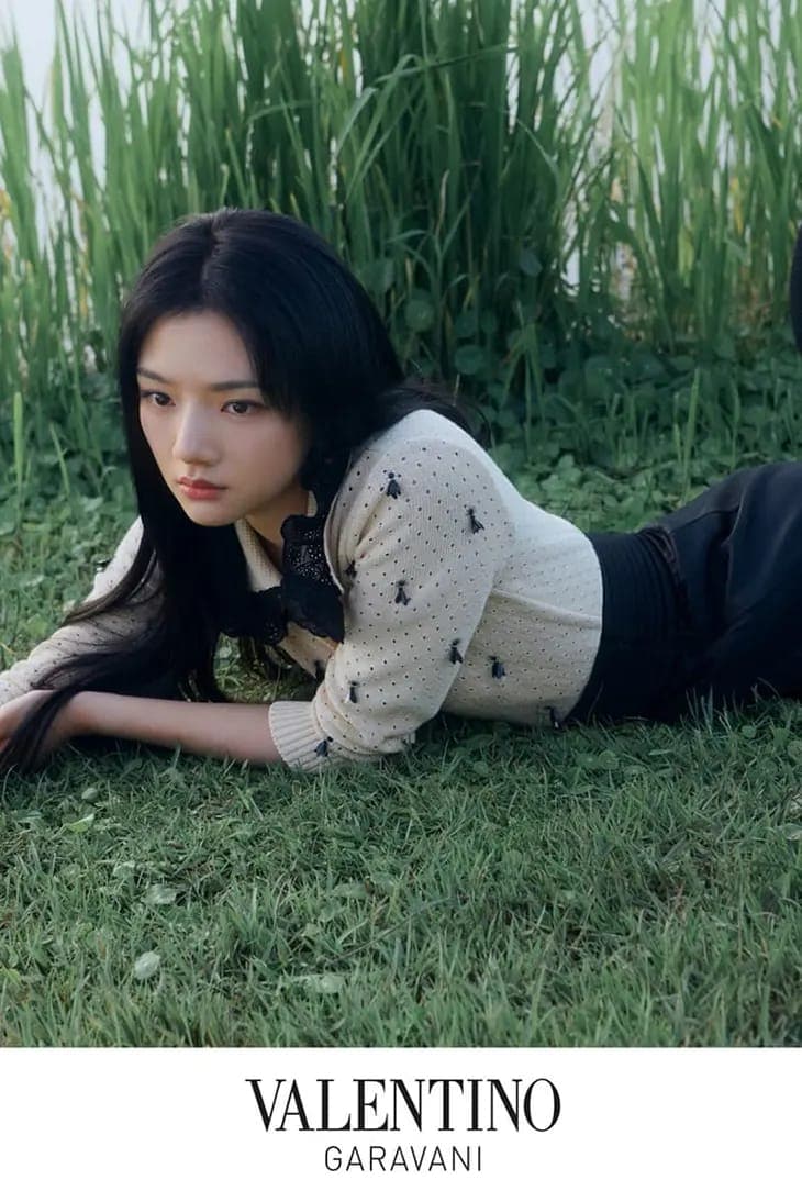 Young woman with long dark hair lying on green grass wearing a cream knitted top, photographed for Valentino.