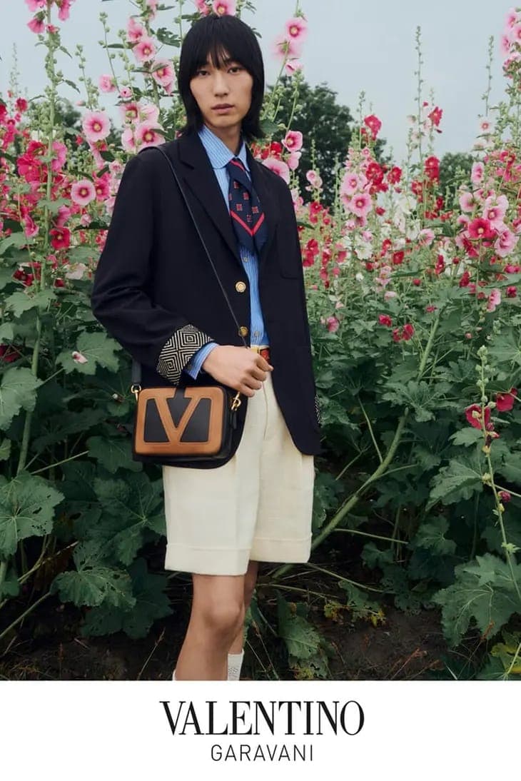 Model standing among tall pink hollyhock flowers wearing a navy blazer, light blue button-down shirt, necktie, and cream shorts with a crossbody handbag.