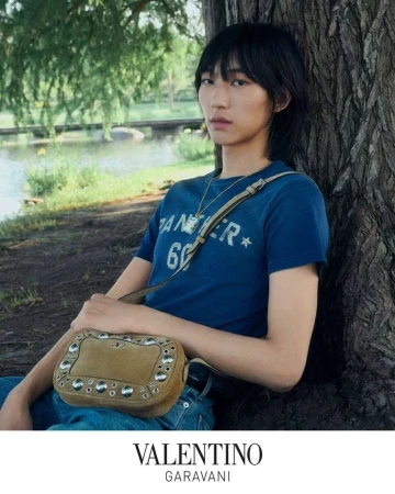 Model leaning against a tree wearing a blue graphic t-shirt and holding a studded tan shoulder bag outdoors by a river.