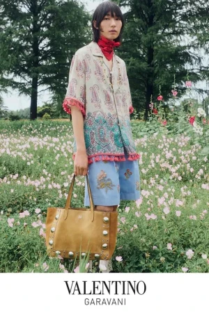 Man standing in a wildflower field holding a tan suede tote bag, wearing a patterned shirt and blue shorts.