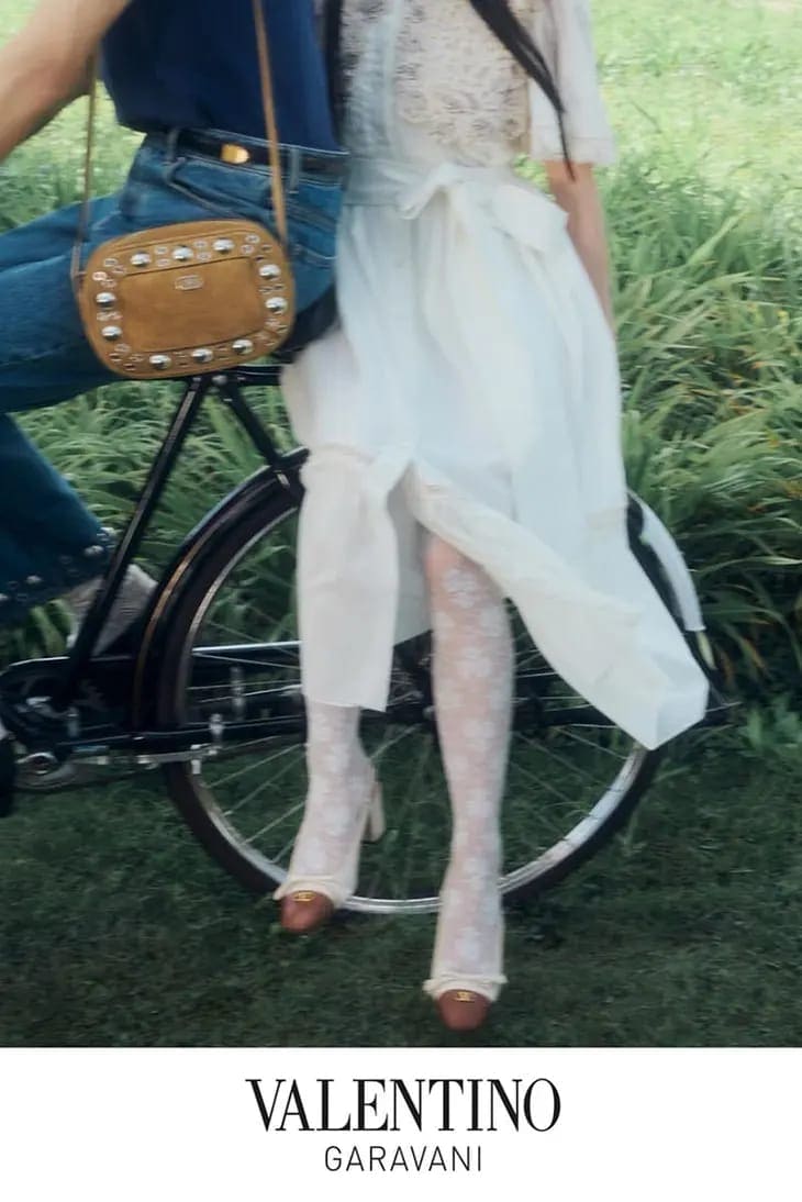 Close-up of two models on a vintage bicycle outdoors in a grassy field wearing Valentino garments and accessories.