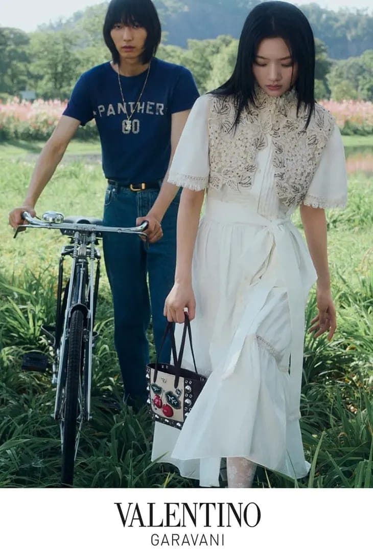 A man holding a vintage bicycle and a woman in a white dress walking through a green meadow for Valentino.