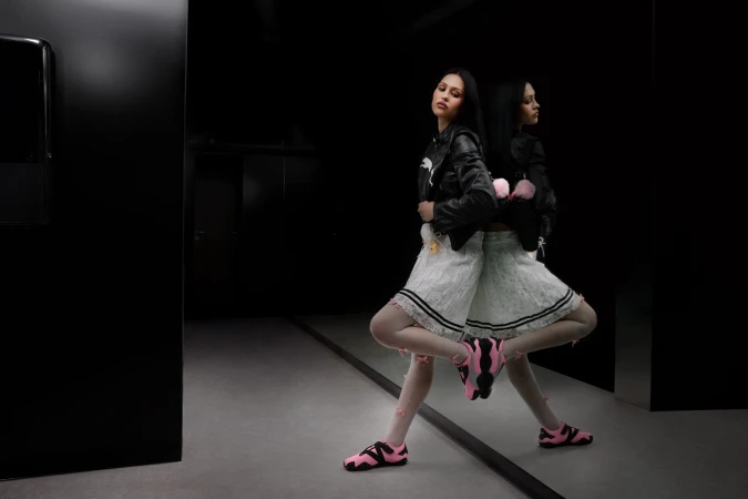 Model Amelia Gray in a black leather jacket and white skirt with pink sneakers posing in a dark interior reflecting in a large mirror