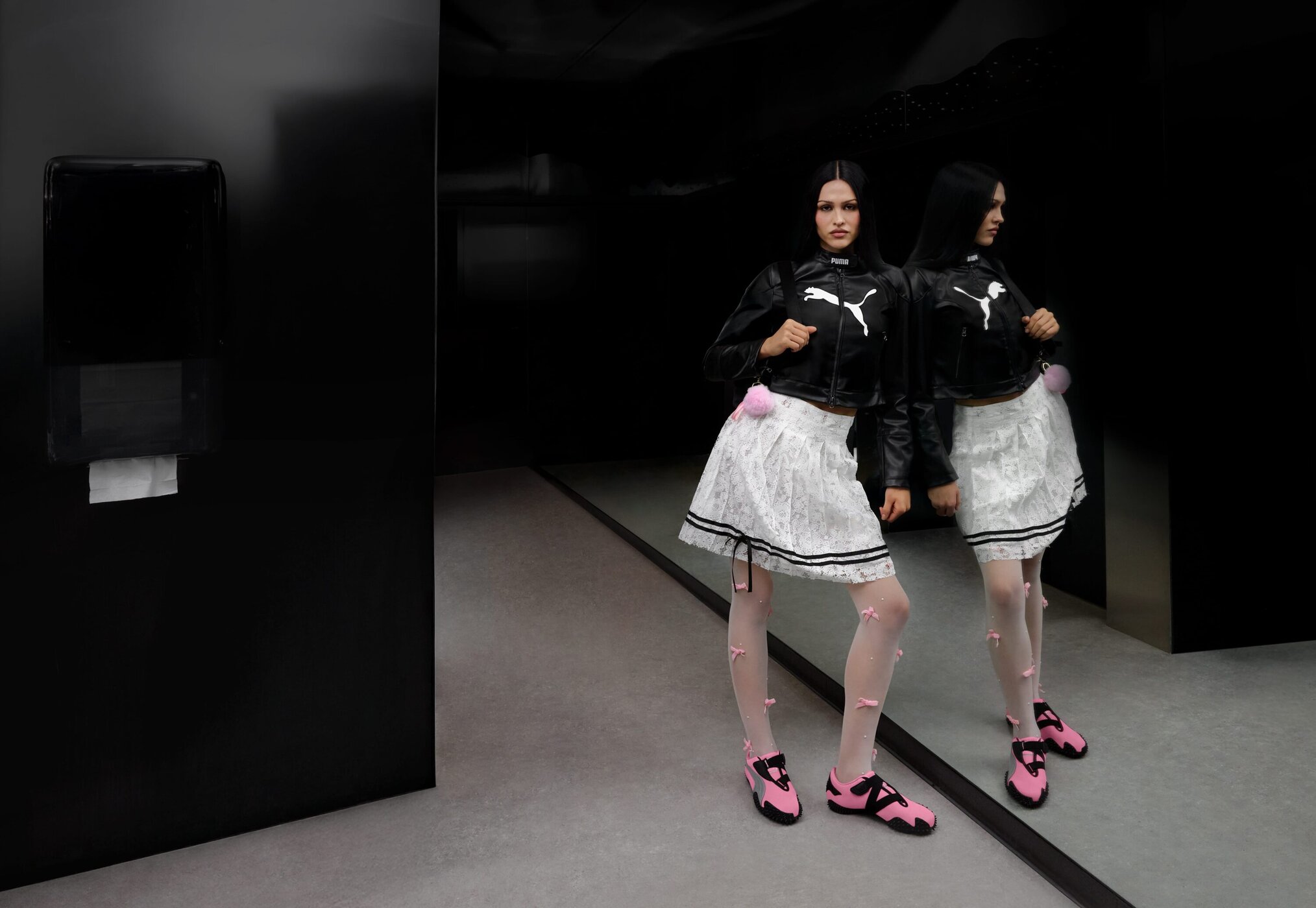 Model Amelia Gray wearing Puma black leather jacket, white lace skirt, and pink sneakers standing before a dark mirror.