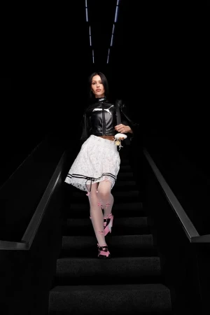 Amelia Gray descending dark stairs wearing a black leather jacket, white lace skirt, and pink sneakers