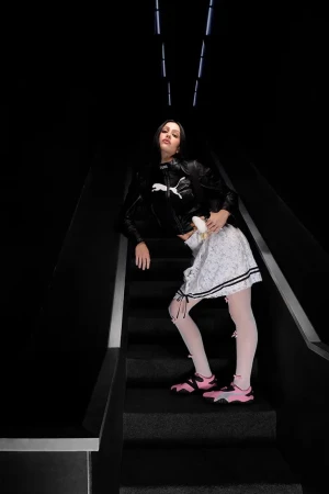 Amelia Gray in a black jacket and white skirt posing on a dark staircase for the Puma Mostro Ad Campaign.