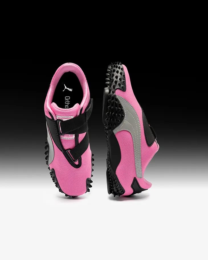Product shot of Puma Mostro sneakers in pink and black viewed from top and side against a gradient background.