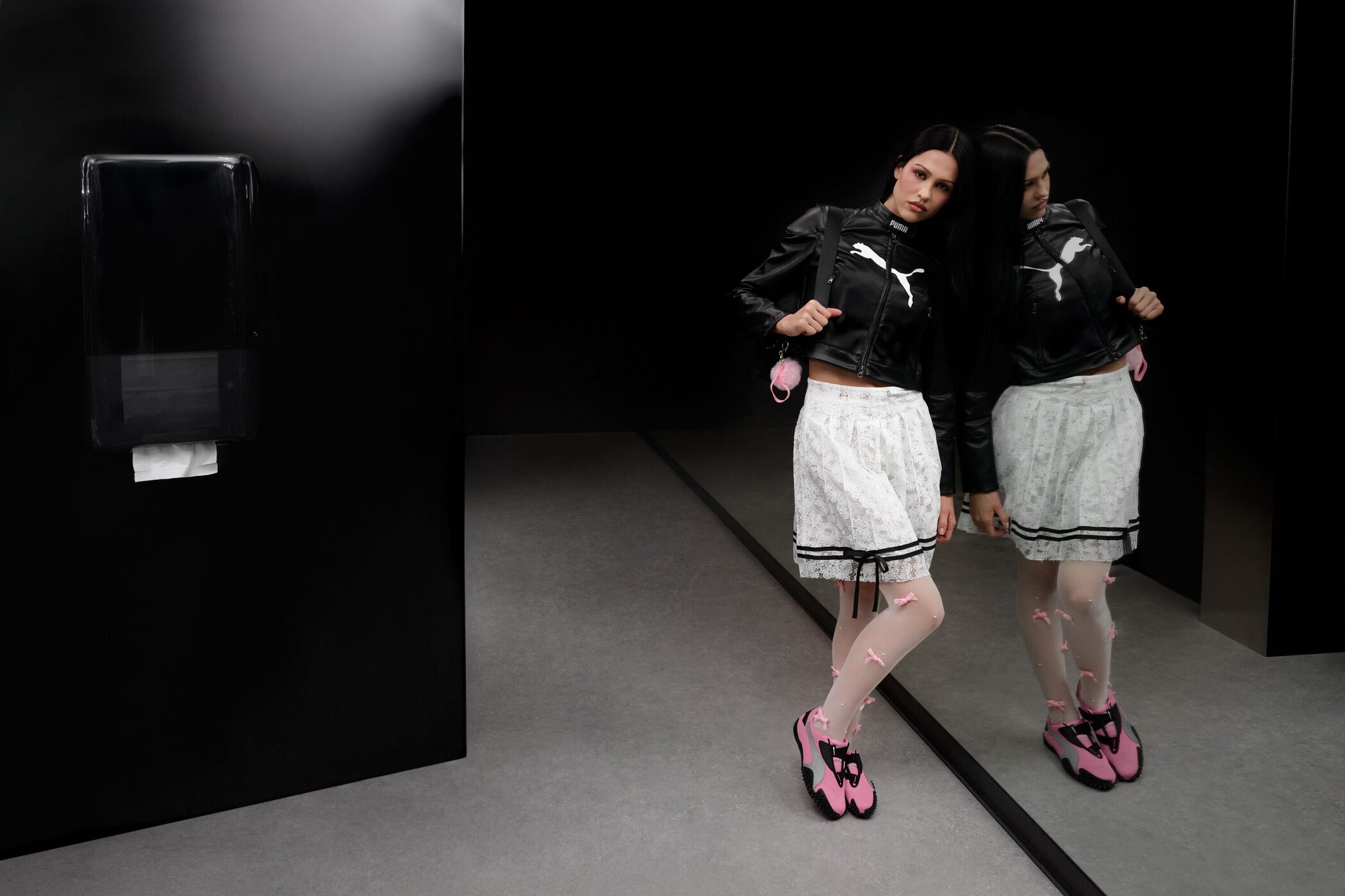 Model Amelia Gray in a black Puma leather jacket and white skirt standing near a dark reflective surface for the Mostro Ad Campaign.
