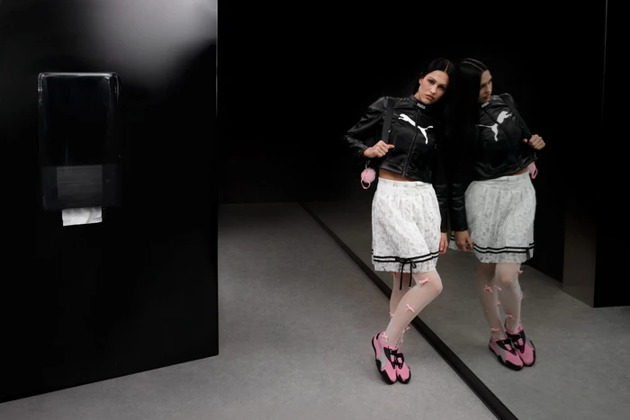 Model Amelia Gray in a black Puma leather jacket and white skirt standing near a dark reflective surface for the Mostro Ad Campaign.
