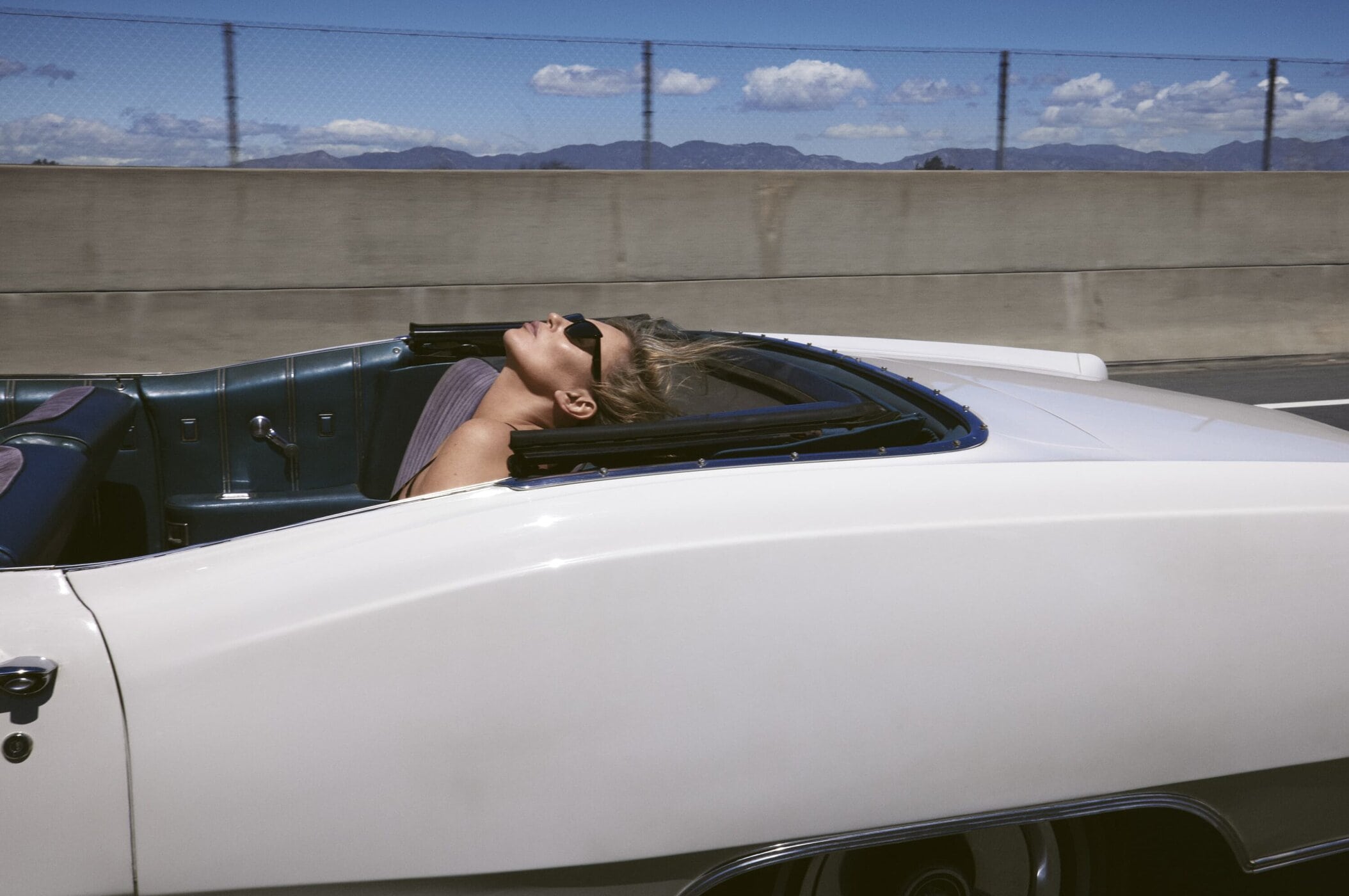 Kate Moss wearing sunglasses leaning back in the passenger seat of a white convertible car on a highway