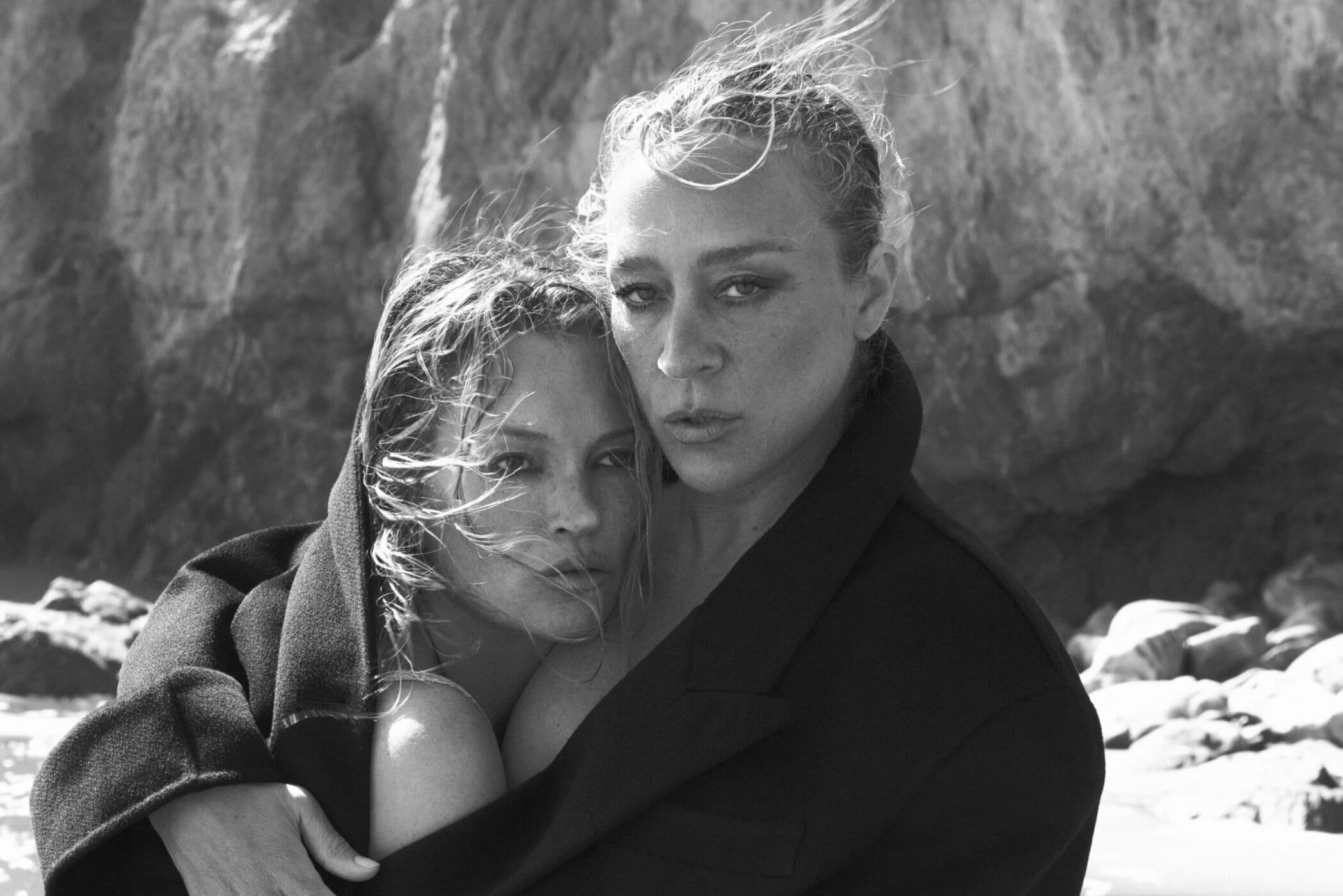 Monochrome portrait of Kate Moss and Chloë Sevigny embracing outdoors against a rocky background for Saint Laurent Pre-Fall 2025