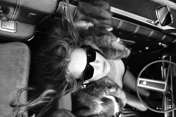 Overhead black and white portrait of Kate Moss wearing sunglasses and fur coat seated in a convertible car