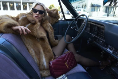 Kate Moss wearing a fur coat and sunglasses seated in a vintage car interior with a burgundy handbag