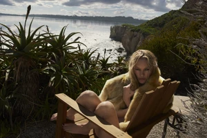 Kate Moss seated in a wooden chair outdoors near a coastal cliff wearing a textured yellow wrap