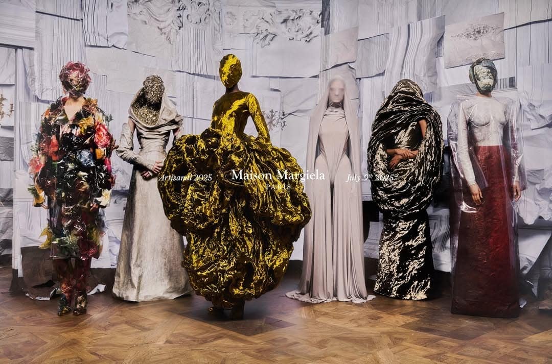 Six models in avant-garde sculptural garments and face coverings standing against a paper-tiled wall