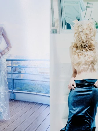 Model wearing a dark skirt and ornate textured top standing on white stairs next to a balcony overlooking a city.