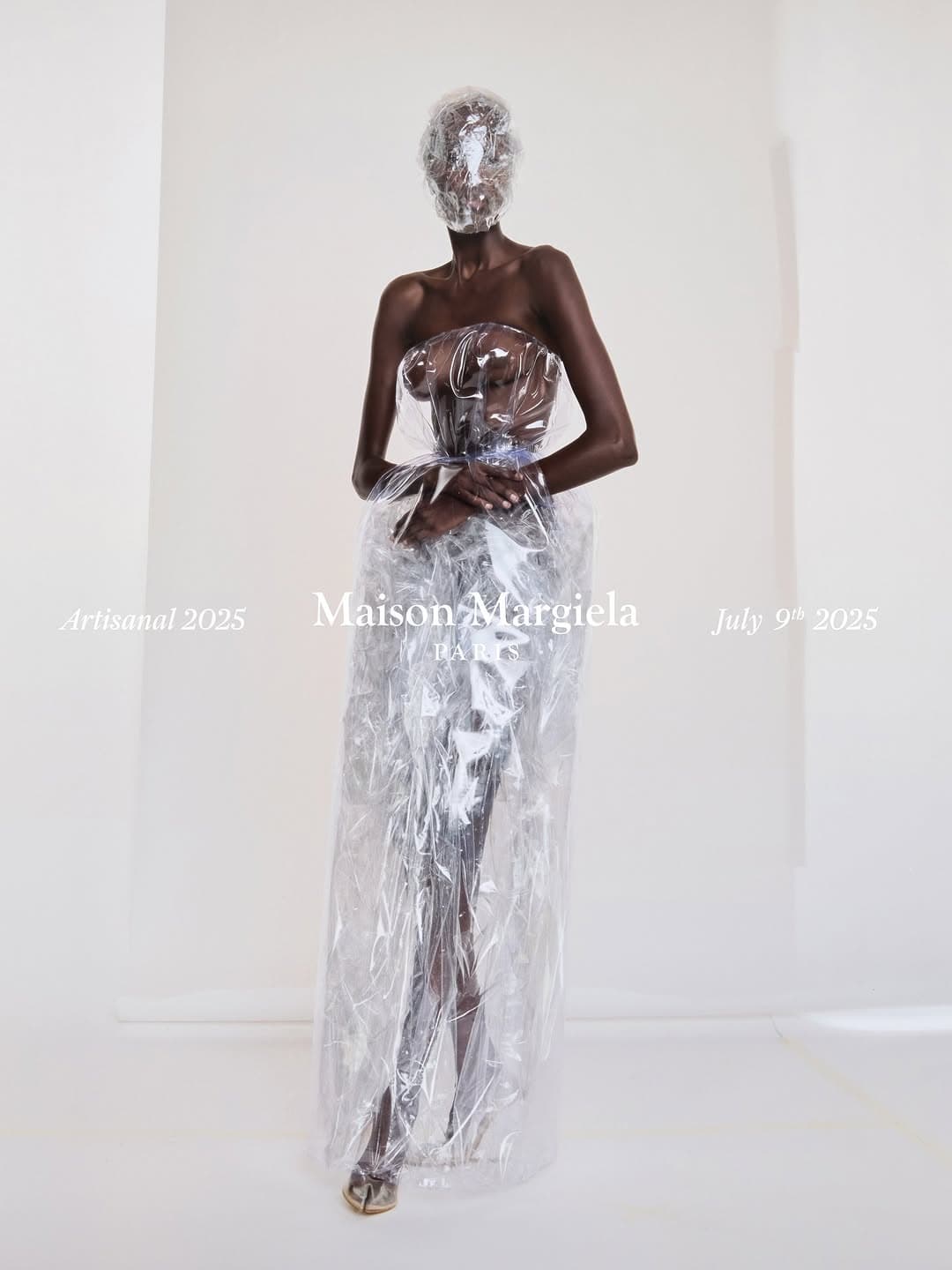 Model wearing a translucent plastic wrapped gown and face covering against a plain white studio background