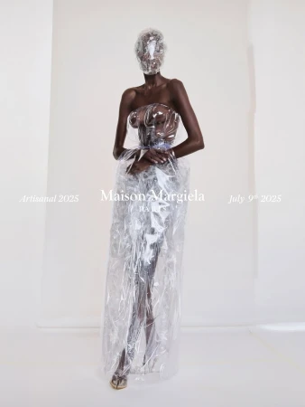 Model wearing a translucent plastic wrapped gown and face covering against a plain white studio background