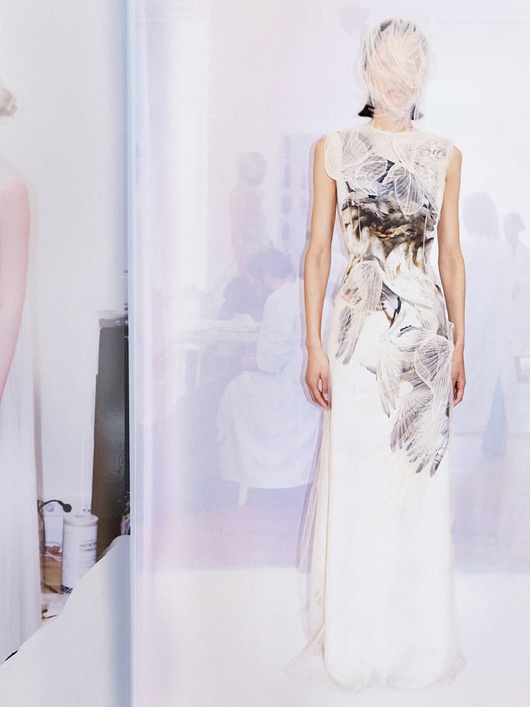 Model in a printed white dress with face covered by a sheer veil standing against a pale, softly lit studio backdrop.