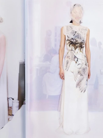 Model in a printed white dress with face covered by a sheer veil standing against a pale, softly lit studio backdrop.